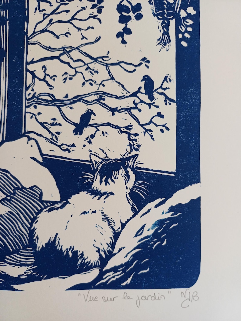 May include: A blue and white linocut print depicting a cat sitting on a surface, looking out a window at two birds perched on tree branches. The print features the text "Vue sur le jardin" and initials at the bottom, rendered in a high-contrast style.