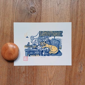 Peut inclure: Une gravure sur bois intitulée "Tea Time" représente un chat se reposant près d'une table avec une tasse à thé. L'impression est en nuances de bleu et de jaune, avec un bouchon en bois à gauche. L'impression a un tampon rouge et la signature de l'artiste.