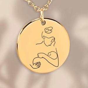 Könnte beinhalten: Goldfarbene Halskette mit einem runden Anhänger. Der Anhänger zeigt eine schwarze Strichzeichnung einer Mutter, die ein Baby hält. Die goldene Kette und der Anhänger bilden ein schlichtes, elegantes Design. Der Anhänger zeigt eine Mutter, die ein Baby hält.