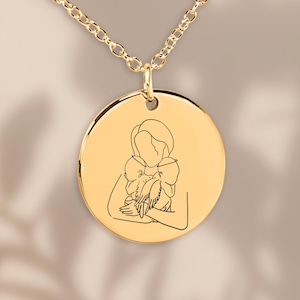 Può includere: Collana d'oro con un ciondolo rotondo che presenta un disegno a linea di una madre che tiene un bambino.