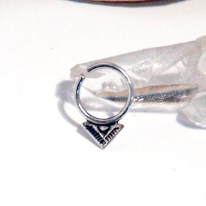 Op de afbeelding: Een zilveren neusring met een geometrische hanger. De ring is cirkelvormig met een kleine, decoratieve driehoekige hanger. De sieraden worden gepresenteerd op een wit oppervlak met een heldere kristal en een stuk hout op de achtergrond.