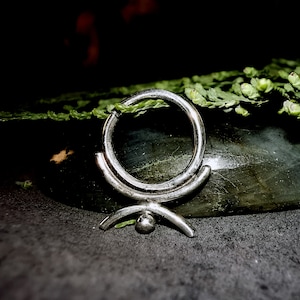 Op de afbeelding: Een zilveren septumring met een uniek ontwerp. De ring heeft een cirkelvormige band met een decoratief element eronder, bestaande uit gebogen lijnen en een kleine bol. De sieraden worden gepresenteerd tegen een donkere achtergrond met groen gebladerte.