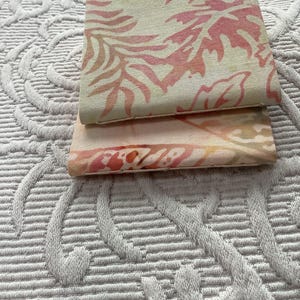 May include: Two small, rectangular fabric items with a leaf pattern in shades of pink and peach. The items are stacked on top of each other, with the top one slightly angled. The background is a white, textured surface.