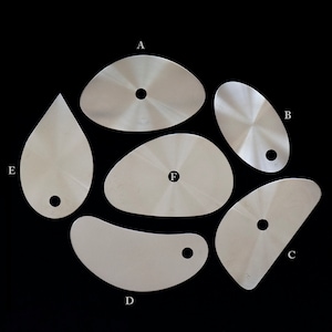 May include: Six different shaped metal palette knives with holes in the center. The knives are arranged in a circle on a black background. The letters A through F are labeled on the knives.