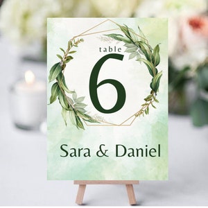 Puede incluir: Una tarjeta con el número de mesa para una boda con un fondo de acuarela en tonos verdes. El número 6 es de color verde oscuro, enmarcado por una forma geométrica dorada y hojas verdes. Los nombres "Sara & Daniel" están impresos en la parte inferior.