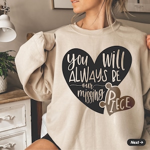 May include: A beige sweatshirt with a black heart design that says "You will always be our missing piece". The heart is a puzzle piece with a smaller heart-shaped piece missing.