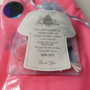 May include: A white card with the text "Washing Instructions" printed on it. The card is inside a pink and white mesh bag. The instructions include: Turn garment inside out, Machine wash cold, Do not use fabric softener, Do not bleach, Tumble Dry Low or hang to dry, Do not iron the design, Do not dry clean. The card also includes a thank you message.