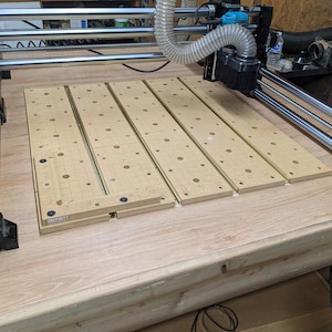 May include: A close-up of a CNC machine with a wooden work surface. The work surface is covered with a grid of holes and has a metal track running along the edge. The machine has a black and silver body with a clear plastic hose attached to it.