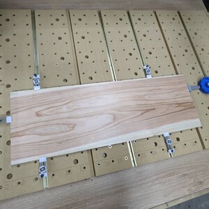 May include: A light wood board is clamped to a workbench with four clamps. The board has a natural wood grain pattern. The workbench has a grid of holes and a ruler along the side.