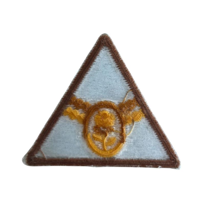 May include: A brown triangular patch with a white background and a yellow embroidered design of a stylized flower with wings.