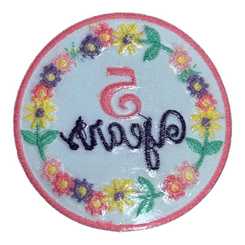 May include: A round, embroidered patch with a white center, featuring the number "5" in pink and the word "Anniversary" in dark blue. The design is encircled by a floral wreath with pink, yellow, and purple flowers, and a pink border.