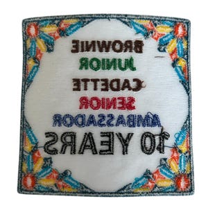 May include: A white embroidered patch with a colorful border. The patch says "10 YEARS" in black, "AMBASSADOR" in blue, "SENIOR" in red, "CADETTE" in brown, "JUNIOR" in green, and "BROWNIE" in brown.