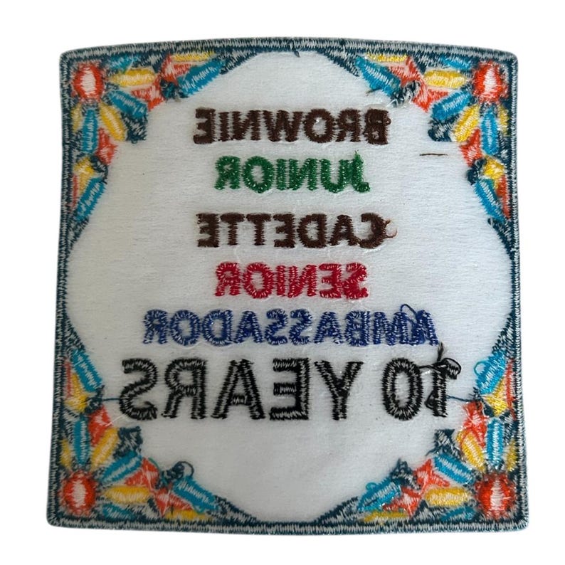 May include: A white embroidered patch with a colorful border. The patch says "10 YEARS" in black, "AMBASSADOR" in blue, "SENIOR" in red, "CADETTE" in brown, "JUNIOR" in green, and "BROWNIE" in brown.