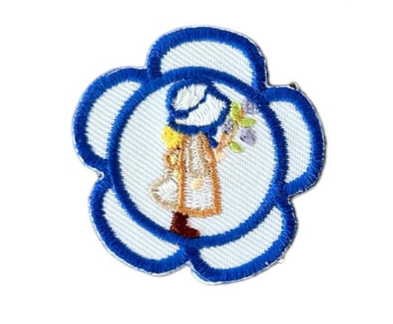 May include: A blue and white embroidered flower patch with a girl in a blue hat and dress holding a bouquet of flowers.