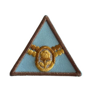 May include: A light blue triangular Girl Scout patch with a gold embroidered design of a rose in an oval frame surrounded by a wreath.
