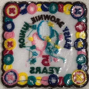 K&ouml;nnte beinhalten: Ein wei&szlig;er gestickter Aufn&auml;her mit braunem Rand. Der Aufn&auml;her zeigt zwei rosa und blaue Ballons mit einem gr&uuml;nen Band und dem Text "Happy Birthday Brownie 5 Years". Der Rand ist mit bunten Kn&ouml;pfen verziert.