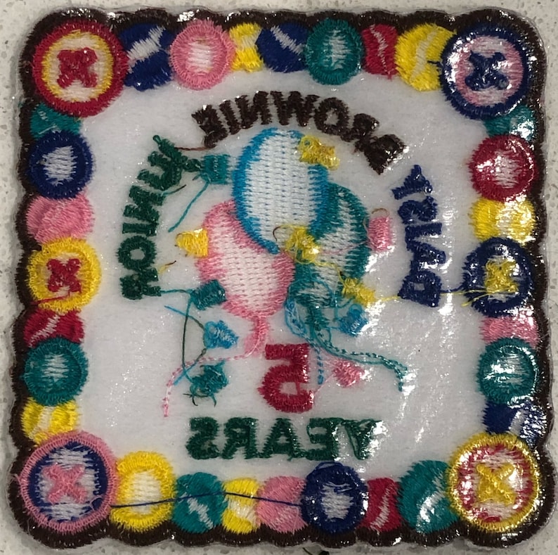 K&ouml;nnte beinhalten: Ein wei&szlig;er gestickter Aufn&auml;her mit braunem Rand. Der Aufn&auml;her zeigt zwei rosa und blaue Ballons mit einem gr&uuml;nen Band und dem Text "Happy Birthday Brownie 5 Years". Der Rand ist mit bunten Kn&ouml;pfen verziert.