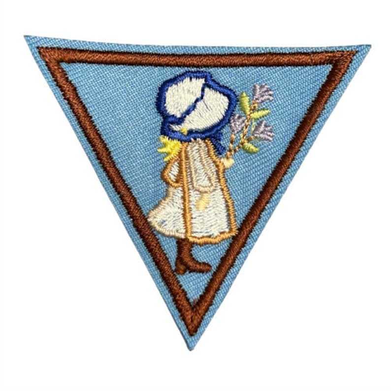 May include: A blue triangular Girl Scout patch with a brown border. The patch features an embroidered image of a girl in a white dress and blue bonnet holding a bouquet of flowers.