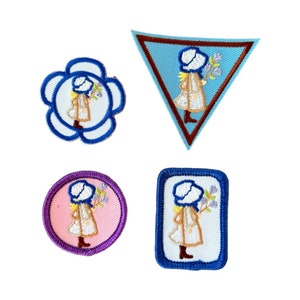 May include: Four embroidered patches featuring a girl in a blue dress and bonnet holding flowers. The patches are in different shapes and colors, including blue, pink, and white.