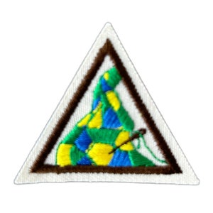 May include: A brown triangular Girl Scout patch with a green, blue, and yellow woven design inside. The design is of a hat with a needle and thread.
