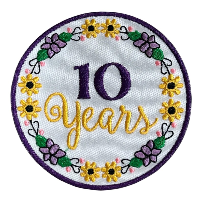 May include: Embroidered patch with a white background, featuring a purple border and the text "10 Years" in purple and gold. The design includes a floral wreath with yellow sunflowers, purple flowers, and green leaves.