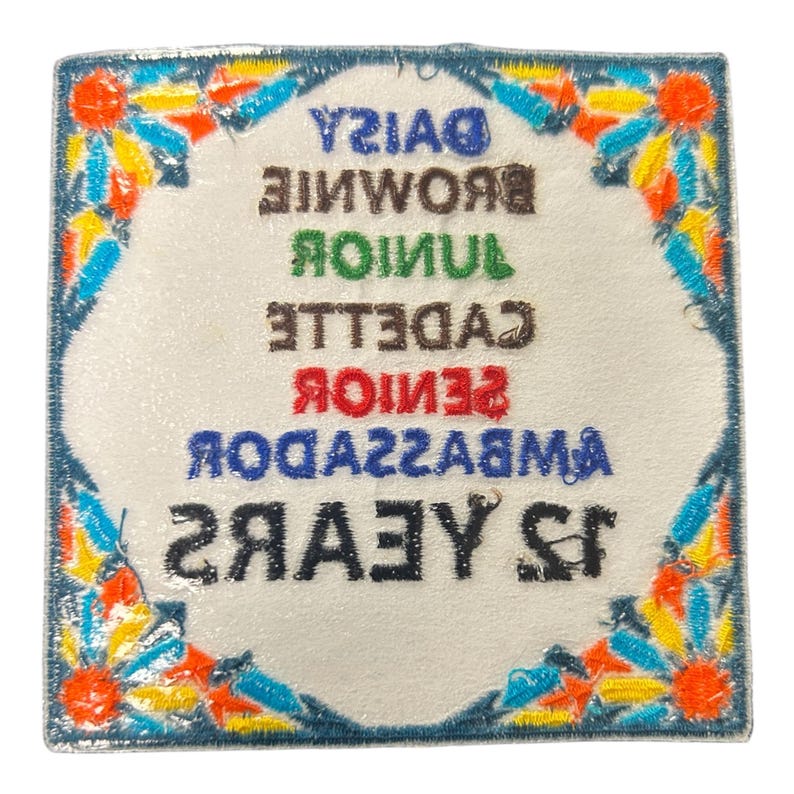 May include: Square embroidered patch with a white center and a blue border. The patch features colorful floral accents and text that reads "DAISY, BROWNIE, JUNIOR, CADETTE, SENIOR, AMBASSADOR, 15 YEARS".