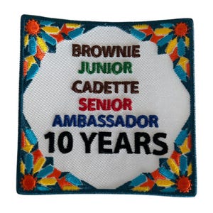 May include: White embroidered patch with a colorful border. The patch says "Brownie, Junior, Cadette, Senior, Ambassador, 10 Years".