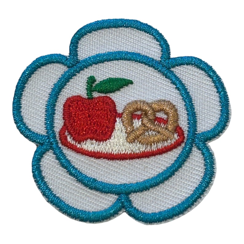 May include: A white flower-shaped embroidered patch with a blue outline. The center of the patch features a red apple and a pretzel on a white plate with a red rim.