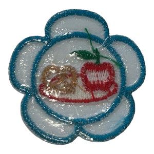 May include: A white flower-shaped patch with a blue outline. The center of the patch features a red apple with a green stem and leaf, and a brown pretzel on a white plate.