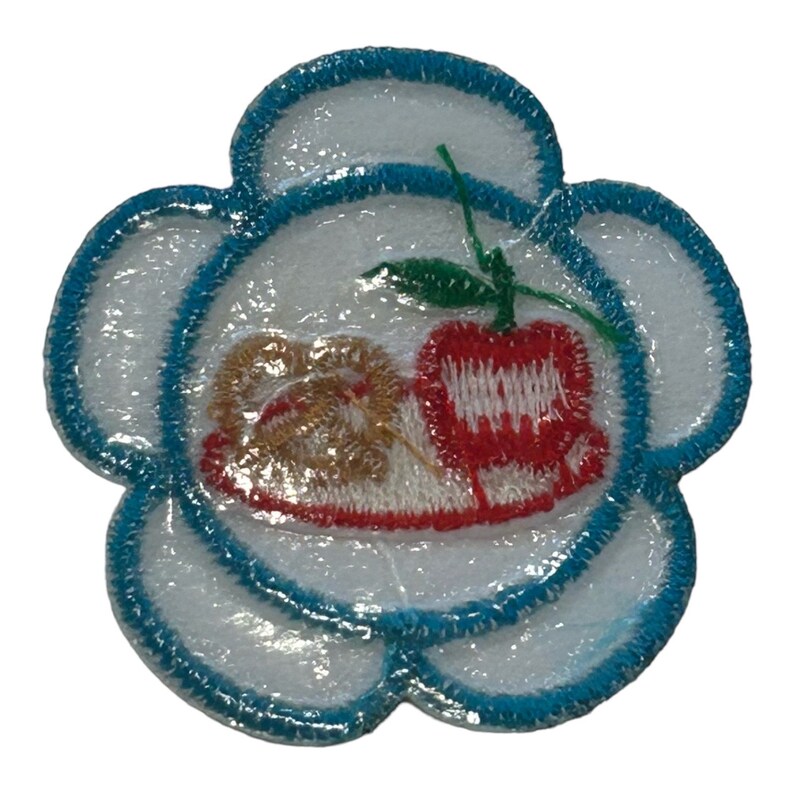 May include: A white flower-shaped patch with a blue outline. The center of the patch features a red apple with a green stem and leaf, and a brown pretzel on a white plate.