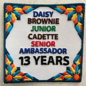 May include: A white patch with a colorful border. The patch has the text "Daisy Brownie Junior Cadette Senior Ambassador 13 Years" in black and colored text.