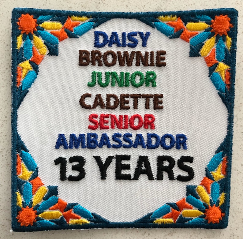 May include: A white patch with a colorful border. The patch has the text "Daisy Brownie Junior Cadette Senior Ambassador 13 Years" in black and colored text.