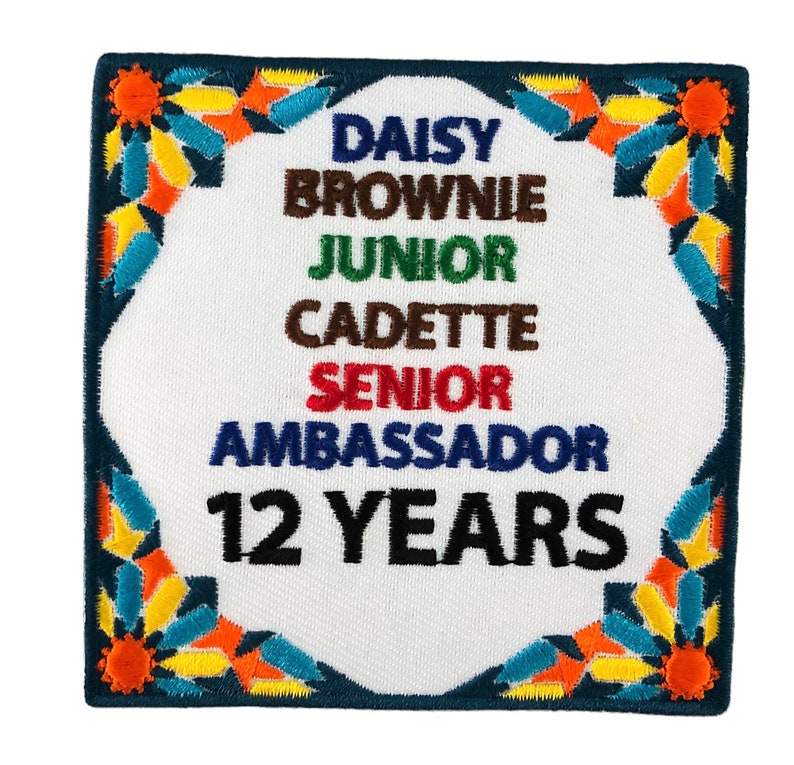 May include: Square embroidered patch with a white center and a dark blue border. The patch features the words "DAISY, BROWNIE, JUNIOR, CADETTE, SENIOR, AMBASSADOR" in various colors, and "12 YEARS" in black. The border has colorful floral accents.