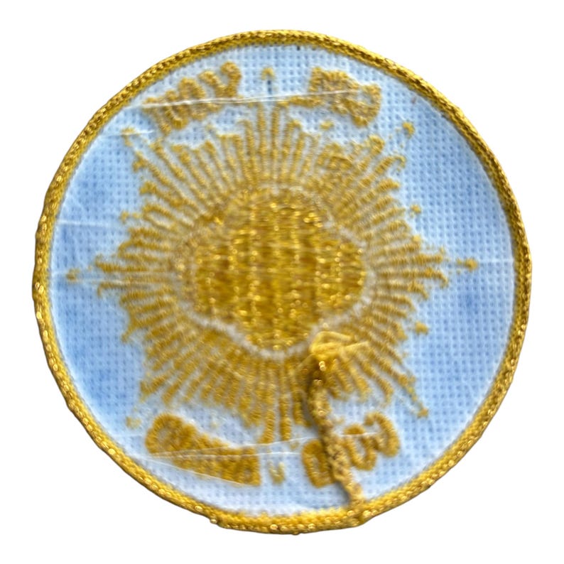 May include: A round embroidered patch with a light blue background and a gold sunburst design. The patch is edged with gold trim and features gold embroidered details.