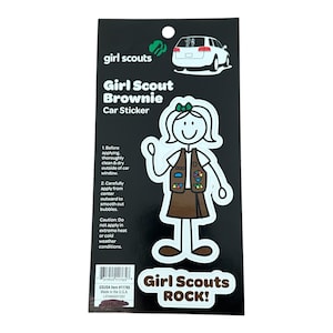 May include: A white car sticker featuring a stick figure of a Girl Scout Brownie wearing a brown uniform and a green sash with badges. The text "Girl Scouts ROCK!" is printed below the figure.