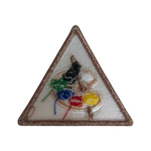 Op de afbeelding: Een driehoekige patch met een bruine rand. Het midden van de patch is wit en heeft een geschilderd palet met vijf kleuren verf: groen, blauw, rood, geel en zwart.