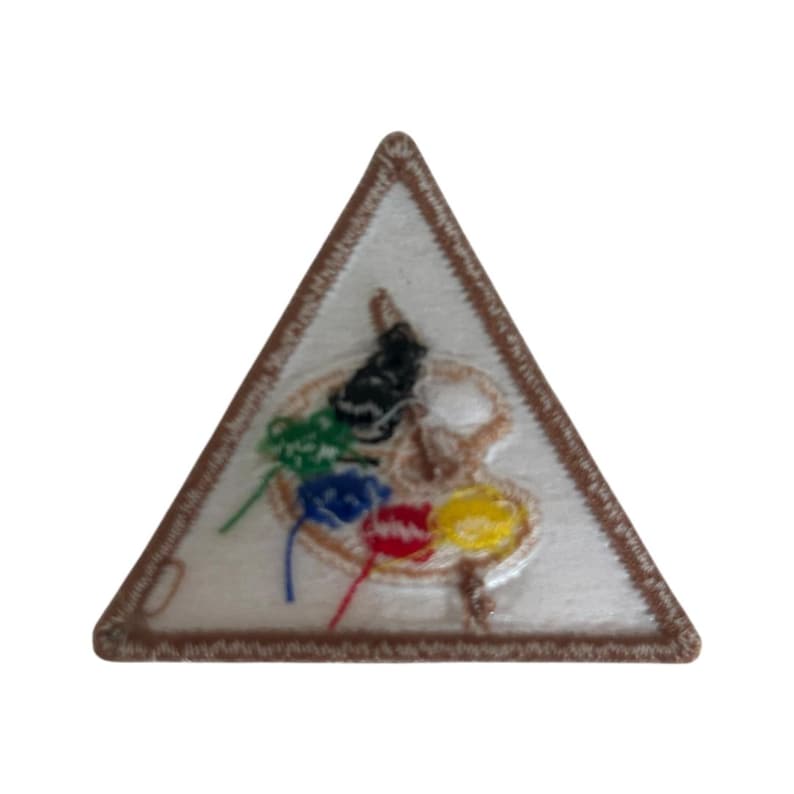 Op de afbeelding: Een driehoekige patch met een bruine rand. Het midden van de patch is wit en heeft een geschilderd palet met vijf kleuren verf: groen, blauw, rood, geel en zwart.