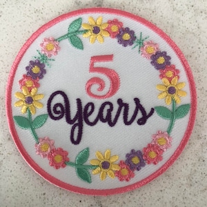 May include: A white embroidered patch with a pink border. The patch features a floral wreath with pink, yellow, and purple flowers. The center of the patch has the number 5 in pink and the text "years" in purple.