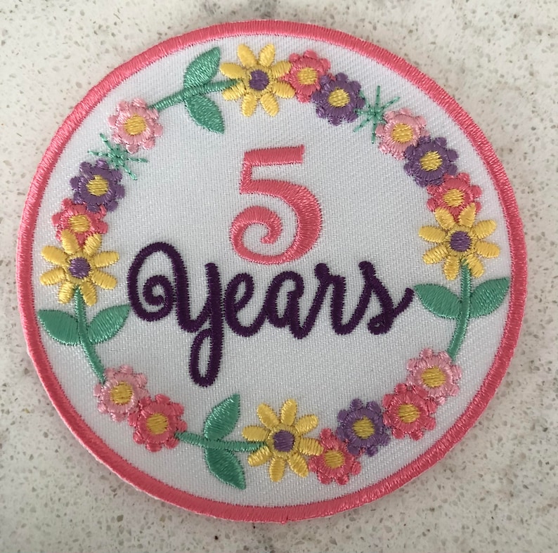 May include: A round, embroidered patch with a white background, featuring the number "5" in pink and the word "years" in purple script. The design is encircled by a wreath of colorful flowers in yellow, pink, and purple, with green leaves, and a pink border.