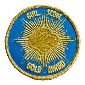 May include: A round, blue Girl Scout Gold Award patch with gold embroidered details. The patch features the words "GIRL SCOUT GOLD AWARD" in gold thread, surrounding a gold sunburst design. The patch has a gold border.