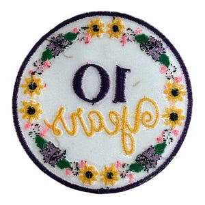 May include: A circular embroidered patch with a white background, a purple border, and a floral wreath design. The patch features the text "OI" in purple and "Kraafs" in yellow cursive.