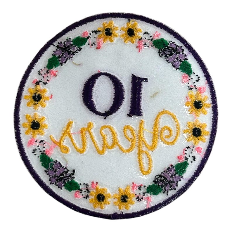 May include: A circular embroidered patch with a white background, a purple border, and a floral wreath design. The patch features the text "OI" in purple and "Kraafs" in yellow cursive.