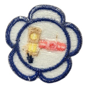 Op de afbeelding: Een witte, bloemvormige patch met een blauwe rand. In het midden van de patch is een gele en bruine illustratie van een persoon die een rood en wit object vasthoudt.