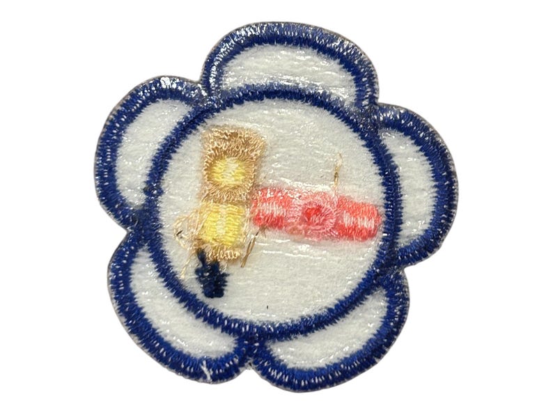 Op de afbeelding: Een witte, bloemvormige patch met een blauwe rand. In het midden van de patch is een gele en bruine illustratie van een persoon die een rood en wit object vasthoudt.