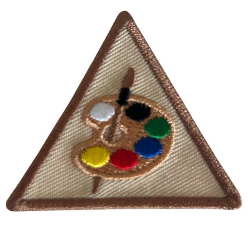 Op de afbeelding: Een driehoekige Girl Scout-badge met een bruine rand. De badge heeft een palet met een kwast, en de kleuren op het palet zijn wit, zwart, geel, rood, blauw en groen.