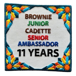 May include: A white embroidered patch with a blue, green, red, and brown border. The patch says "Brownie, Junior, Cadette, Senior, Ambassador, 11 Years".