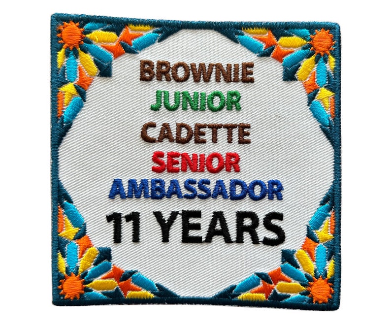 May include: A white embroidered patch with a blue, green, red, and brown border. The patch says "Brownie, Junior, Cadette, Senior, Ambassador, 11 Years".
