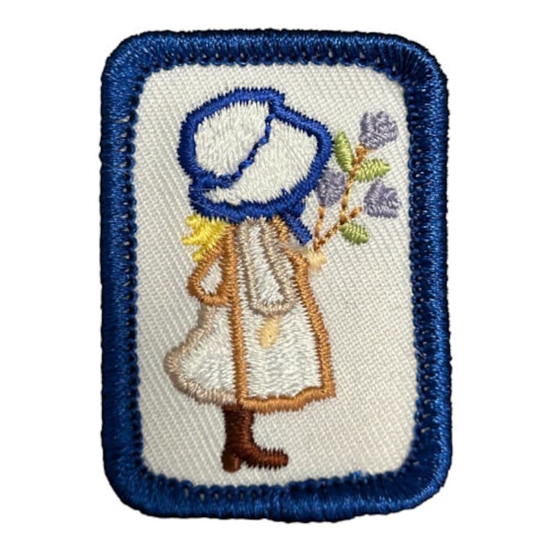 May include: An embroidered patch featuring a girl in a blue bonnet and white dress holding a bouquet of purple flowers. The patch is surrounded by a blue border.
