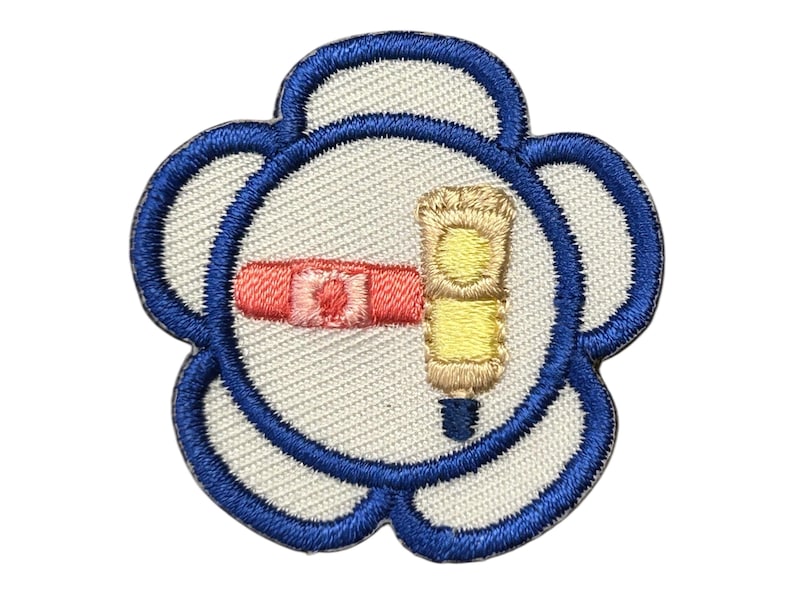 Op de afbeelding: Een geborduurde bloemenpatch in blauw en wit met een roze en gele tube handcr&egrave;me erin.