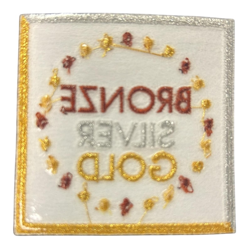 May include: Square patch with the words "BRONZE", "SILVER", and "GOLD" in glittery lettering. The patch has a gold border and a silver border. A decorative wreath of gold and red accents surrounds the words.